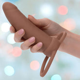 A hand with light pink nail polish holds the CalExotics Performance Maxx Rechargeable Thick Dual Penetrator in brown, featuring dual rings at the base, against a blurred pastel bokeh background.